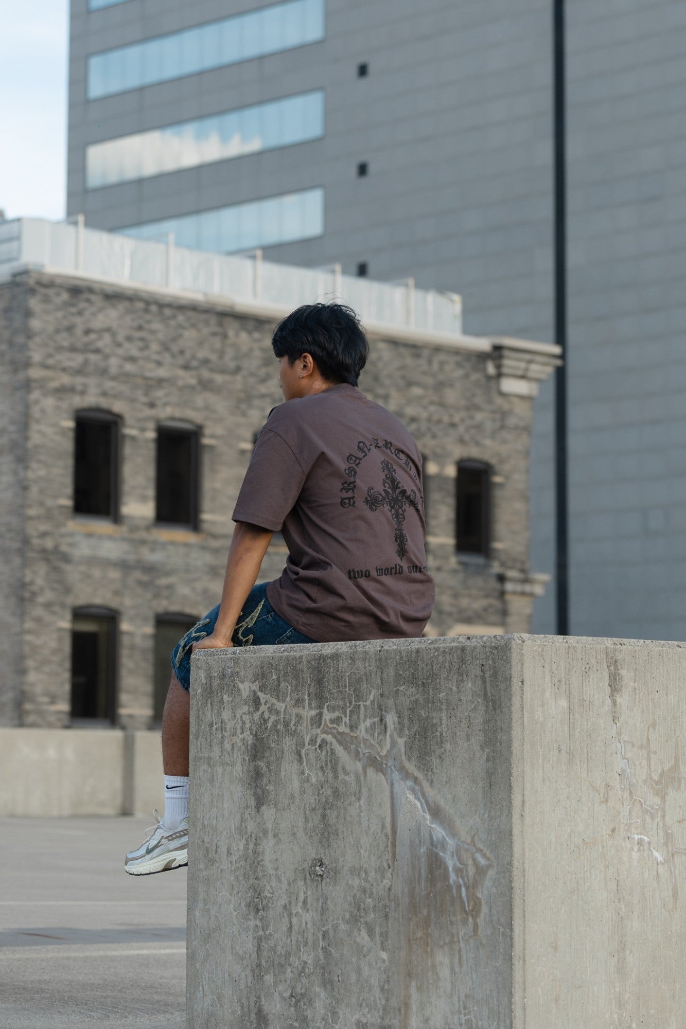 Sitting on a rooftop ledge in the brown Arsan Archive oversized tee, back print showing
